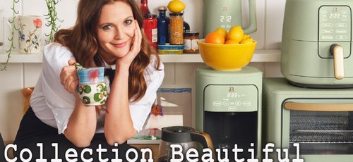 Drew Barrymore sourit en tenant une tasse colorée dans une cuisine au style moderne. Elle est entourée d'appareils électroménagers vert sauge de sa "Collection Beautiful", incluant une friteuse à air, une cafetière, une bouilloire et un grille-pain four. Le décor est lumineux et chaleureux, avec des touches de plantes et d'aliments frais, créant une ambiance conviviale.